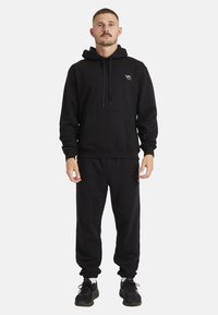 Sort hættetrøje med snøre og frontlomme, parret med matchende sorte sweatpants. Har et lille logo på hættetrøjen.