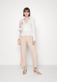 Blusa branca com tecido texturizado, combinada com calças largas bege e sapatos de salto castanhos claros. Os acessórios incluem uma bolsa de palha.