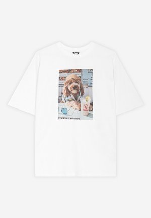 T-shirt blanc avec un motif d’un chien portant un chapeau et une chemise, assis à une table avec des lunettes de soleil et une limonade avec une paille.