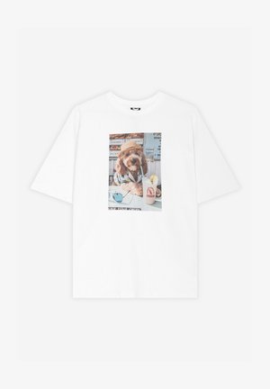 T-shirt blanc avec un motif d’un chien portant un chapeau et une chemise, assis à une table avec des lunettes de soleil et une limonade avec une paille.