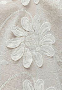 Motif floral brodé blanc avec de grandes pétales et un fond de tissu texturé aux tons beige clair.