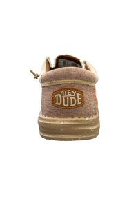 Zapatilla de lona color cáñamo con un parche de cuero marrón etiquetado "HEY DUDE", cordones beige en contraste y una suela de goma texturada con manchas.
