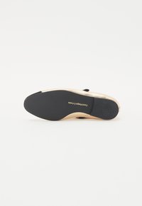 Zapato de suela plana con parte superior de cuero dorado, acentos negros y suela de goma texturizada, con el nombre de la marca estampado en la parte inferior.