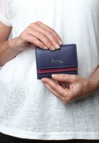 Navy leather wallet with a red accent at the fold, featuring a snap closure and embossed logo, held in a hand against a light background.