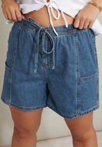 Pantalones cortos de mezclilla azul con cintura elástica, cierre con cordón y dos grandes bolsillos delanteros; diseño casual y de corte relajado.