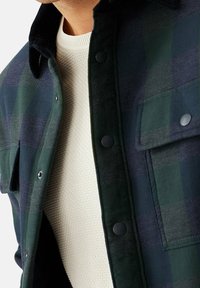 Grøn og marineblå ternet skjortejakke med sorte knapper, en kordfløjlskrave og en struktureret cremesweater under. Har brystlommer.