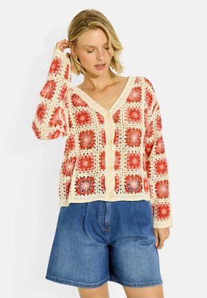 Femme portant un cardigan en crochet crème et rouge avec un motif floral carré et un short en jean bleu, regardant vers le bas avec une main près de la tête.