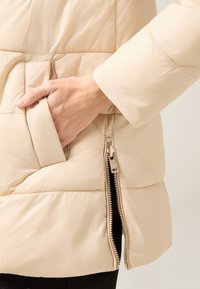 Chaqueta acolchada beige con textura acolchada, bolsillo lateral con cremallera y manga ajustada. El material tiene un acabado liso y ligeramente brillante.