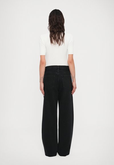 Hosszú sötét hajú személy, fehér rövid ujjú felsőt és magas derekú, bőszárú fekete farmert viselve, háttal állva fehér háttér előtt.