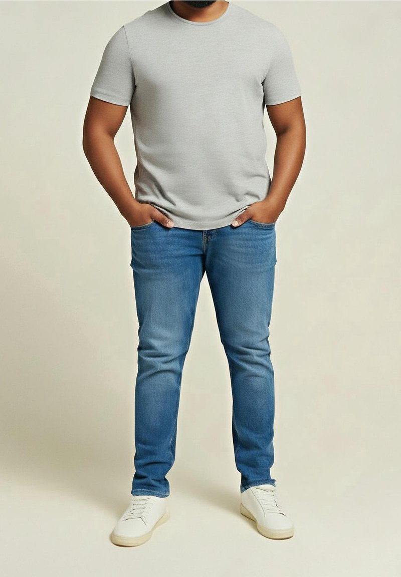Homme portant un t-shirt gris clair à manches courtes, un jean bleu et des baskets blanches, debout les mains dans les poches devant un fond uni.