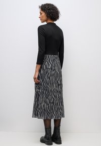 Frau mit lockigem Haar, die ein eng anliegendes schwarzes Oberteil, einen Midi-Rock mit Zebramuster, schwarze Strumpfhosen und schwarze Stiefeletten trägt, und vor einer schlichten Wand steht.