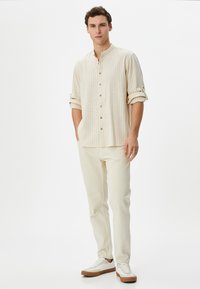 Camisa de manga larga color beige claro con rayas verticales, tapeta de botones y puños enrollables, combinada con pantalones crema y zapatillas blancas con suelas marrones.