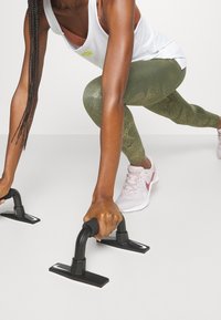 Barre per flessioni nere su una superficie bianca; mani femminili che afferrano i manici. Indossando Leggings a fantasia verde oliva e scarpe rosa.