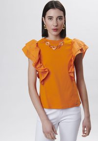 Camiseta naranja con mangas acampanadas, escote redondo y un collar decorativo de cadena. Combinada con pantalones blancos. Textura de algodón suave.