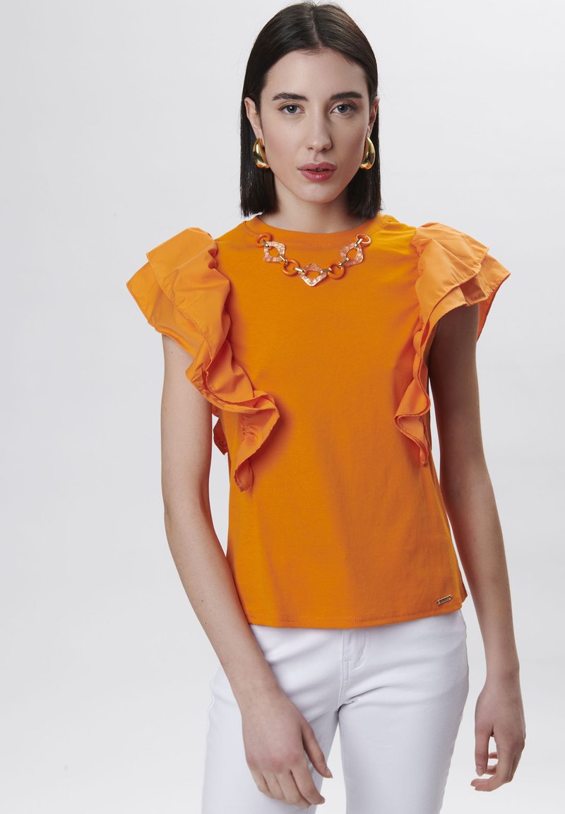Camiseta naranja con mangas acampanadas, escote redondo y un collar decorativo de cadena. Combinada con pantalones blancos. Textura de algodón suave.