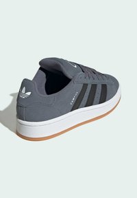Zapatillas de ante gris con diseño de tres franjas negras y suela de goma blanca. Presentan un cuello acolchado y una suela de goma texturizada.