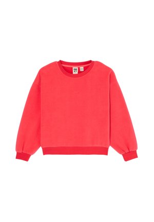 Fille portant un sweat corail, un jean large bleu et des chaussons beiges, souriante avec une main touchant sa tête sur un fond blanc.