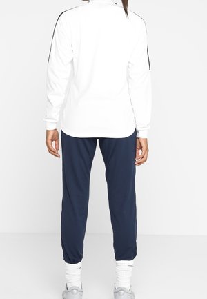 Person steht mit dem Rücken zur Kamera, trägt ein weißes Langarmshirt mit schwarzen Streifen und marineblaue Jogginghose mit weißen Sneakers und Socken.