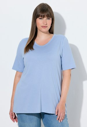 Jonge vrouw met stijlbruin haar, gekleed in een lichtblauw T-shirt met korte mouwen en V-hals en een blauwe spijkerbroek, staand tegen een witte achtergrond.