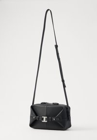 Bolsa de ombro em pele preta com forma retangular, apresentando um design frontal com um detalhe semelhante a um laço e acabamentos em prata. Alça ajustável.