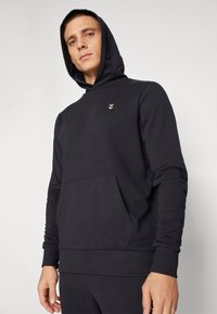 Mężczyzna w czarnej bluzie z kapturem z przednią kieszenią i małym białym logo, stojący na tle jasnego, jednolitego tła.