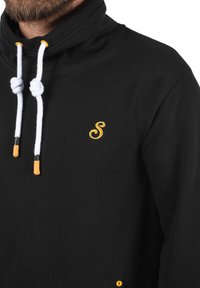 Suéter negro con cuello alto, que presenta cordones blancos con puntas amarillas y un logo amarillo bordado en el pecho.