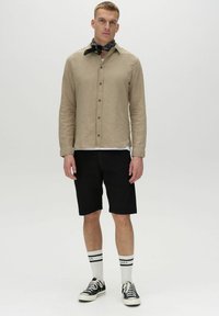Beige Langarmhemd mit Knöpfen, schwarze Shorts, schwarzes Bandana, schwarze Sneakers und weiße Socken mit schwarzen Streifen. Einfaches, lässiges Ensemble.