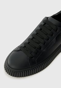 Schwarzer Sneaker mit dickem, geriffelten Sohlen, abgerundeter Spitze und passenden schwarzen Schnürsenkeln auf einem hellen Hintergrund.