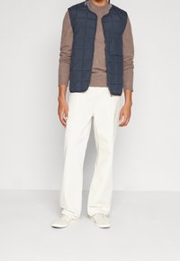 Mörkblå quiltad väst över en brun tröja, ihop med off-white vida byxor och ljusa sneakers, med en ficka med dragkedja på västen.