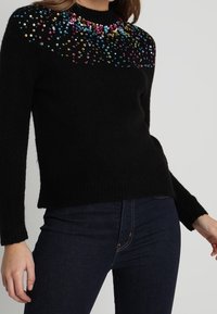 Pull noir présentant un motif décoratif coloré en sequins sur le haut de l'avant. Texture douce avec manches longues et coupe décontractée.