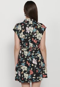 Mujer con cabello largo castaño, vistiendo un vestido negro sin mangas con un estampado floral colorido, de pie de espaldas contra un fondo sencillo.