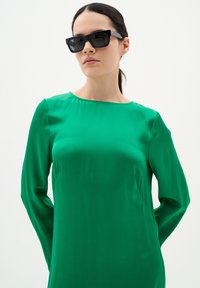 Zöld, hosszú ujjú ruha sima textúrával és kerek nyakkivágással. Fekete, oversized napszemüveg teszi teljessé a megjelenést, egyszínű háttér előtt.