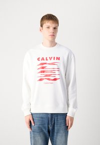 Felpa bianca con grafica rossa "CALVIN" e linee orizzontali, colletto rotondo, polsini a costine e vestibilità rilassata. Indossata con jeans blu.