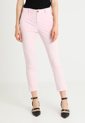 Femme portant un pantalon slim rose clair et des talons noirs à bout pointu avec bride à la cheville, debout devant un fond clair uni.