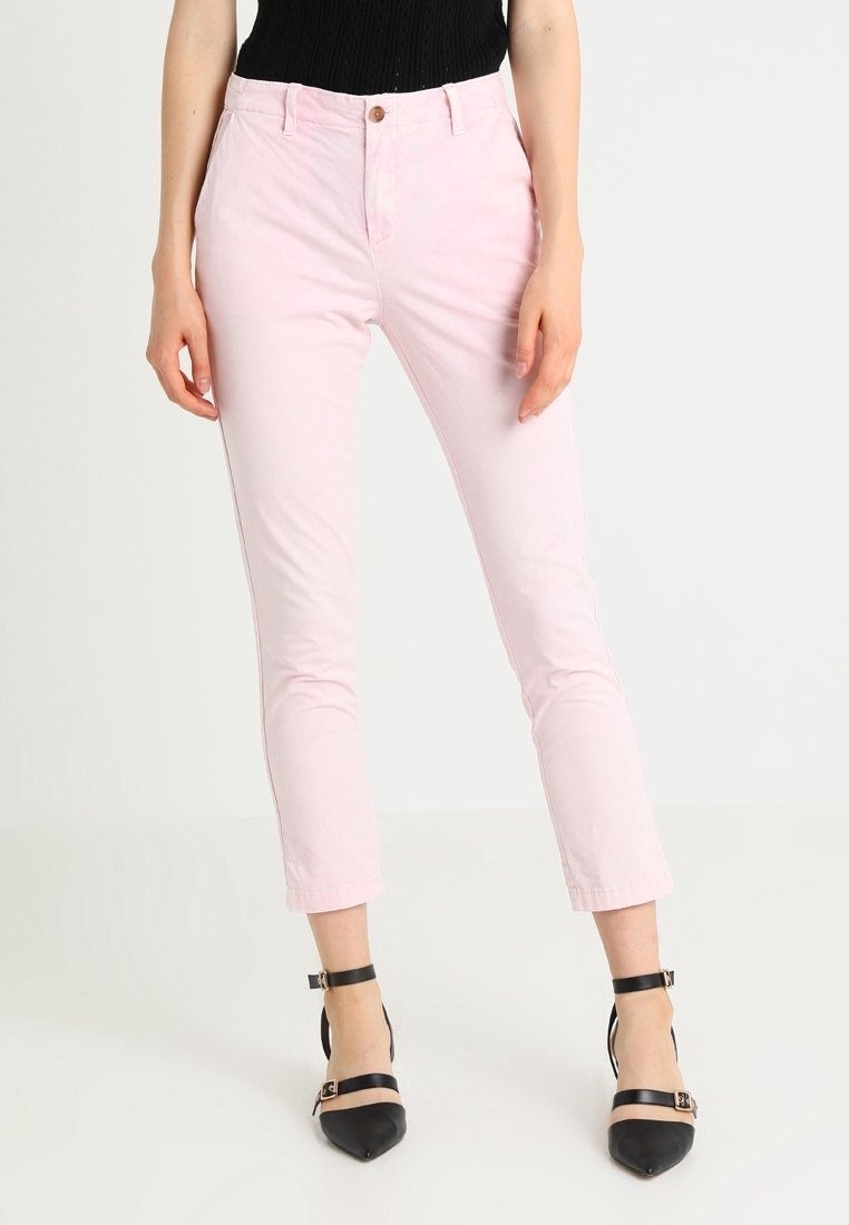 Femme portant un pantalon slim rose clair et des talons noirs à bout pointu avec bride à la cheville, debout devant un fond clair uni.