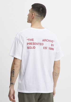 Camiseta de algodón blanca con texto rojo en la parte trasera: “EL ARCHIVO” “PRESENTADO POR SOLID EST. 1999.” Manga corta y corte relajado.