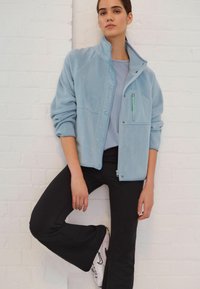 Svijetloplava polar fleece jakna s visokim izrezom, gumbima na pritisak i džepom na prsima sa zatvaračem. Uparena s crnim širokim hlačama i tenisicama.