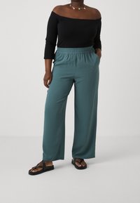 Svart off-shoulder topp och teal vida byxor med elastisk midja, med en mjuk textur. Bärs med svarta flip-flops.