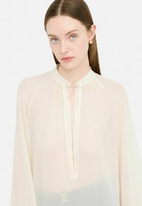 Gennemsigtig cream blouse med høj krave, nøglehulshalser, perleaccentuer og plisserede detaljer. Har lange ærmer og en let tekstur.