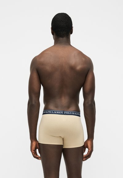 Homme debout de dos, portant un boxer beige avec une ceinture bleu marine marquée "Polo Ralph Lauren" sur un fond blanc uni.
