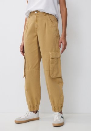 Personne portant un pantalon cargo beige avec des bas élastiques et des baskets blanches sur un fond blanc uni.