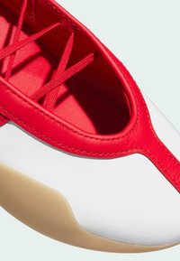 Crvena i bijela sportska cipela s gornjim dijelom od glatke kože, crvenim vezicama i zaobljenim potplatom koji ima gumenu donju površinu i kontrastne šavove.