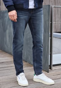 Tummansiniset farkut, joissa on slim fit -leikkaus, hienovaraiset kulumat ja erottuva ompelu, yhdistettynä valkoisiin tennareihin.