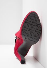 Rode suède hoge hak met een puntige neus, zijrits en een gestructureerde zwarte rubberen zool met het merknaam "Tamaris" duidelijk herhaald.