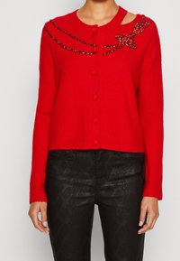 Cardigan rouge en maille avec encolure ronde, fermeture boutonnée à l'avant, et détail orné de pierres noires sur les épaules. Longueur courte.