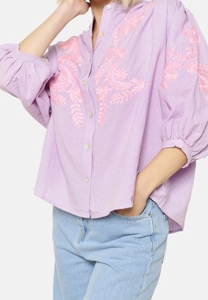 Frau trägt eine lockere lavendelfarbene Bluse mit pinker Blumenstickerei und hellblaue Jeans, Hand in der Tasche, Ellbogen angewinkelt.