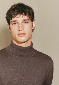 Brauner Rollkragenpullover mit strukturiertem Strick, der einen hohen Kragen und eine figurnahe Passform aufweist. Der Hautton ist hell mit sichtbaren Sommersprossen.