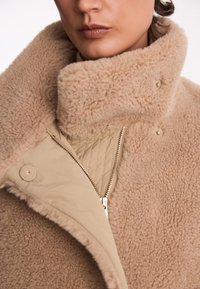 Beżowy płaszcz z owczej wełny z wysokim kołnierzem, miękką teksturą, zatrzaskami i zamkiem błyskawicznym oraz pikowaną podszewką.