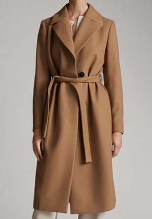 Vrouw die een knie-lange, camelkleurige wollen jas met een ceintuur draagt, met brede revers en een zichtbare knoop, staand tegen een effen achtergrond.