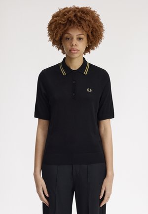 Femme aux cheveux bouclés portant un polo noir à manches courtes avec une bordure dorée sur le col et un pantalon noir, debout devant un fond uni.
